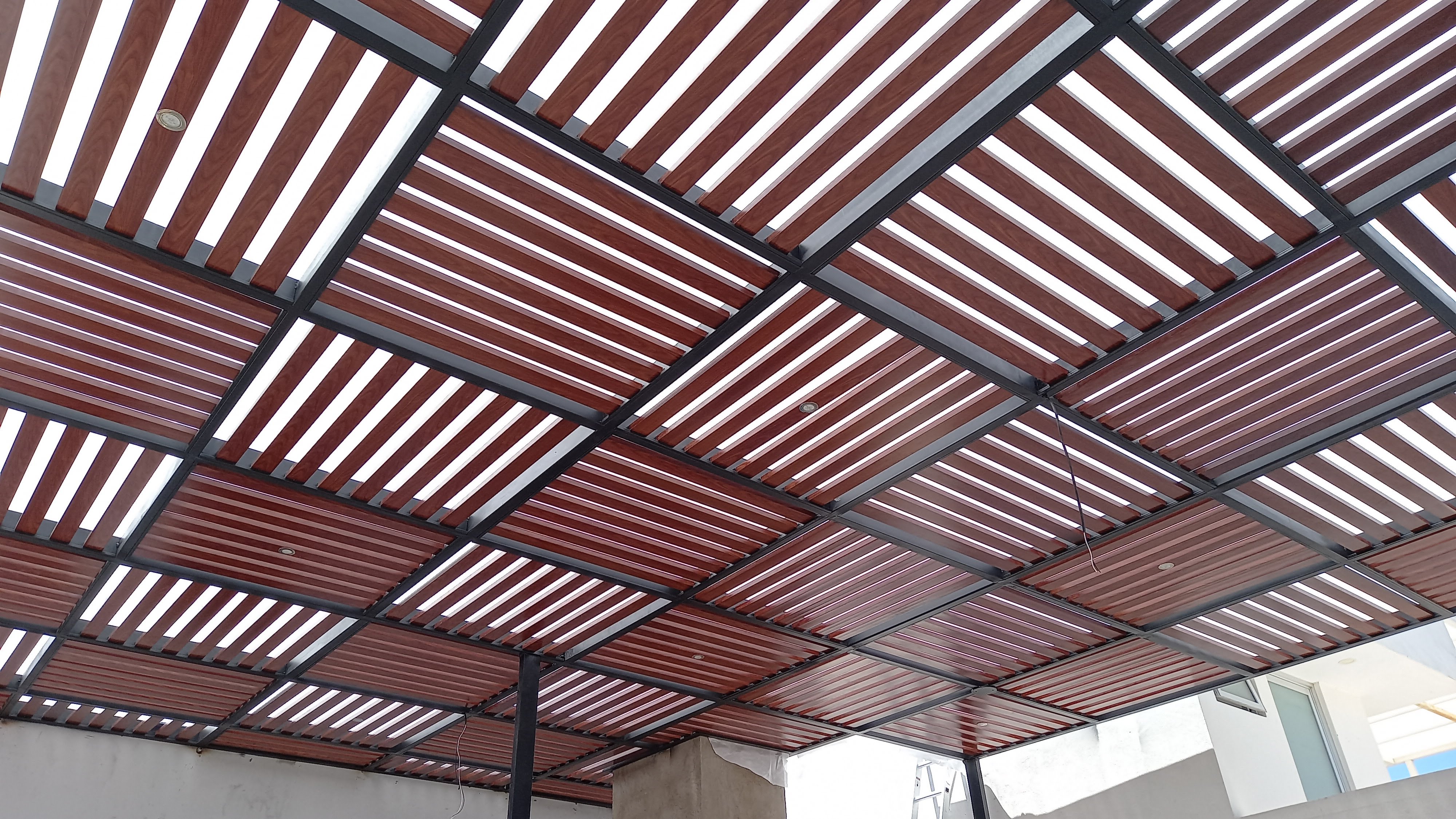 Pérgola moderna de acero adosada al muro, con cubierta tipo tablero de ajedrez que proyecta sombra cuadriculada sobre la terraza
